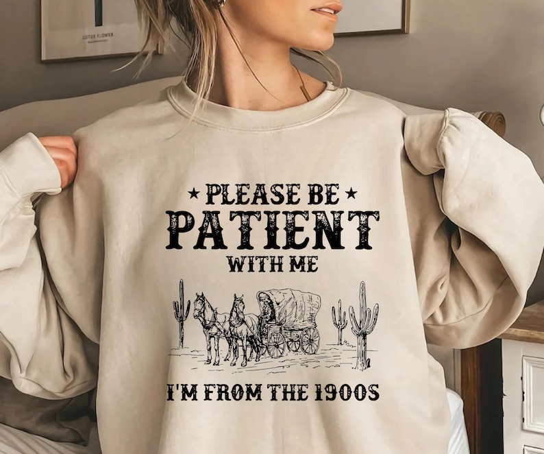 May include: A beige sweatshirt with a black graphic print that says "Please be patient with me I'm from the 1900s". The graphic also includes a black and white illustration of a horse-drawn wagon with cacti.