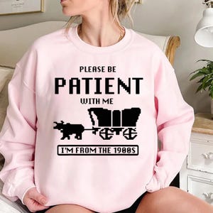 May include: Pink sweatshirt with black text that reads "Please be patient with me I'm from the 1980s". The text is in a pixelated font and there is a pixelated image of a covered wagon pulled by an ox.