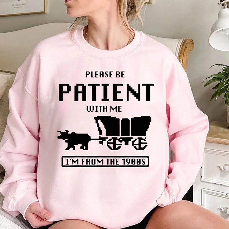 May include: Pink sweatshirt with black text that reads "Please be patient with me I'm from the 1980s". The text is in a pixelated font and there is a pixelated image of a covered wagon pulled by an ox.