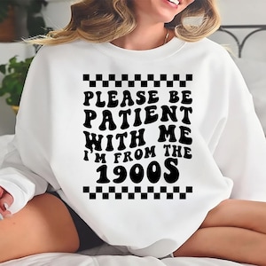 May include: White sweatshirt with black text that reads "Please be patient with me I'm from the 1900s". The text is in a retro font style with a checkered pattern above and below the text.