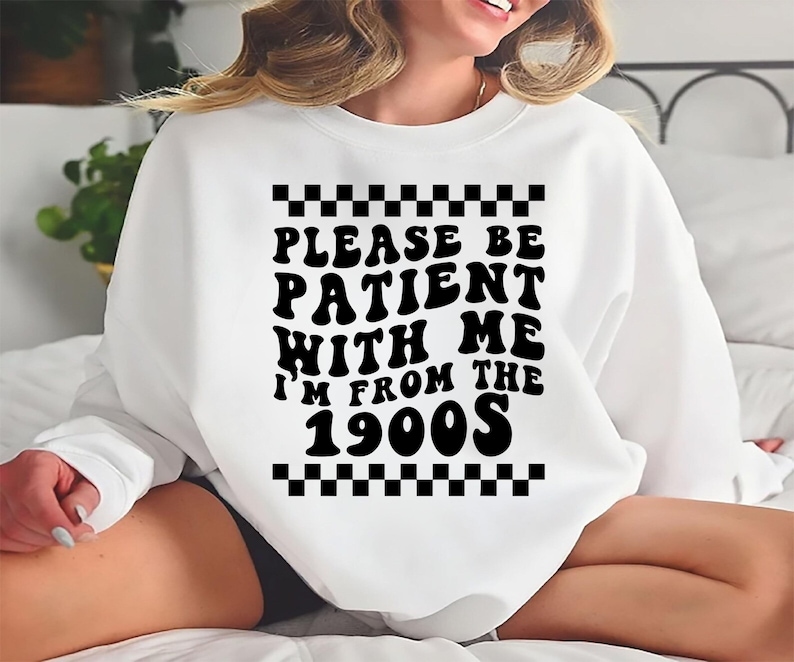 May include: White sweatshirt with black text that reads "Please be patient with me I'm from the 1900s". The text is in a retro font style with a checkered pattern above and below the text.