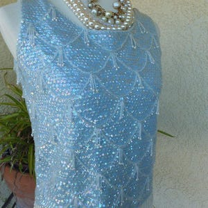 Puede incluir: Una blusa sin mangas de color azul claro cubierta de lentejuelas iridiscentes dispuestas en un patrón de festones. La prenda presenta delicados flecos de cuentas blancas que cuelgan de cada festón y a lo largo del dobladillo inferior.