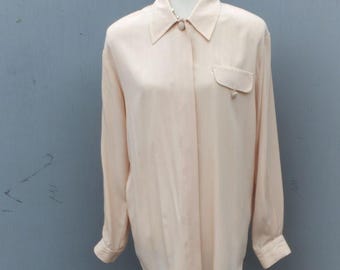 May include: A light peach-colored long-sleeve button-down shirt with a collared neckline and a single chest pocket. The shirt is displayed on a wooden mannequin. The fabric appears to be lightweight and flowing.