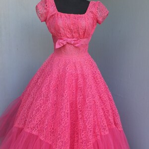 May include: A vintage pink lace and tulle ball gown with a fitted bodice and a full skirt. The dress has a square neckline and short sleeves. A pink satin ribbon is tied around the waist.