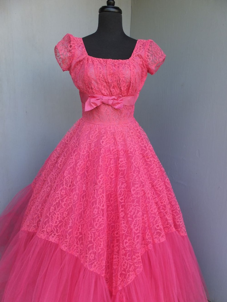 May include: A vintage pink lace and tulle ball gown with a fitted bodice and a full skirt. The dress has a square neckline and short sleeves. A pink satin ribbon is tied around the waist.