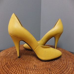 Puede incluir: Un par de zapatos de tacón alto amarillos con punta puntiaguda. Los zapatos son de cuero y tienen un tacón de aguja.