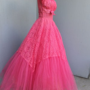 May include: A vintage pink tulle and lace dress with a fitted bodice and a full skirt. The dress has a sweetheart neckline and a bow at the waist.