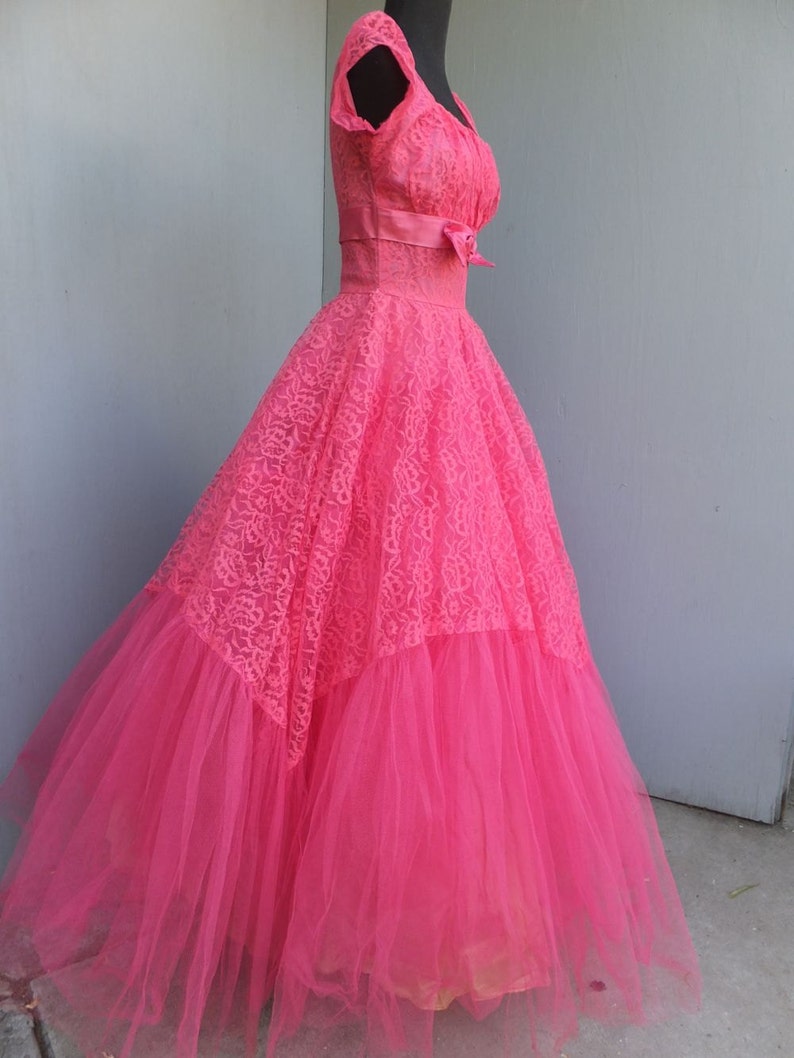 May include: A vintage pink tulle and lace dress with a fitted bodice and a full skirt. The dress has a sweetheart neckline and a bow at the waist.
