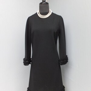 Puede incluir: Un vestido negro de manga larga con un dobladillo con volantes. El vestido tiene un escote redondo y est&aacute; adornado con un collar de perlas.