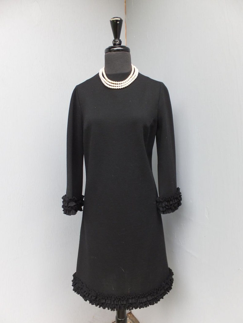 Puede incluir: Un vestido negro de manga larga con un dobladillo con volantes. El vestido tiene un escote redondo y est&aacute; adornado con un collar de perlas.