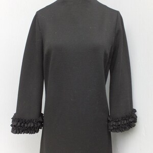 Puede incluir: Un vestido negro con cuello alto y mangas largas. Las mangas tienen un ribete de volantes en los pu&ntilde;os.