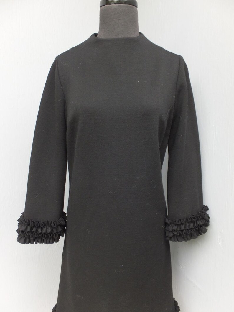 Puede incluir: Un vestido negro con cuello alto y mangas largas. Las mangas tienen un ribete de volantes en los pu&ntilde;os.