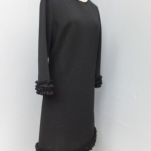 Puede incluir: Un vestido negro con un dobladillo y pu&ntilde;os con volantes. El vestido tiene un escote redondo y mangas largas. Un collar de perlas se lleva en un maniqu&iacute; que lleva el vestido.