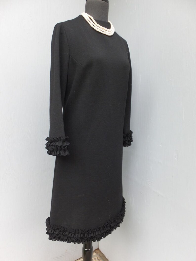 Puede incluir: Un vestido negro con un dobladillo y pu&ntilde;os con volantes. El vestido tiene un escote redondo y mangas largas. Un collar de perlas se lleva en un maniqu&iacute; que lleva el vestido.
