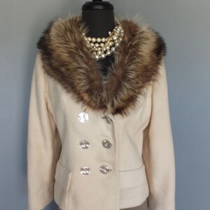 Puede incluir: Una chaqueta de lana de color crema con un gran cuello de piel marrón y cuatro botones plateados. La chaqueta está en un maniquí y tiene una falda marrón debajo.