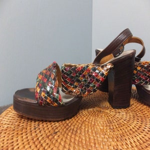 May include: A pair of brown platform sandals with a woven multi-colored strap. The strap is made of leather and has a geometric pattern. The sandals have a chunky heel.