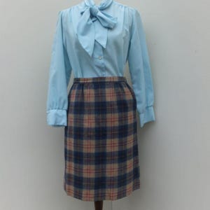 Op de afbeelding: Een vintage outfit met een lichtblauwe blouse met een strikkraag en lange mouwen, gecombineerd met een knielange geruite rok. De rok heeft een patroon van blauwe, beige en rode ruiten. De outfit wordt gepresenteerd op een mannequin.