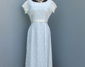 Puede incluir: Un vestido vintage de encaje marfil con cuello redondo y mangas cortas. El vestido presenta un corpiño ajustado, un cinturón de cinta de satén y una falda fluida. Un collar de perlas adorna el cuello del maniquí. El vestido se exhibe en un maniquí negro.