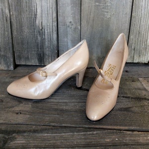 May include: A pair of beige leather pumps with a Mary Jane strap and a gold-tone buckle. The shoes have a pointed toe and a mid-height heel. The brand name "Ferragamo" is visible on the insole. The shoes are displayed on a wooden surface.