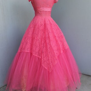 May include: A pink tulle and lace gown with a fitted bodice and a full skirt. The gown has a sweetheart neckline and short sleeves. The bodice is decorated with a pink satin ribbon.