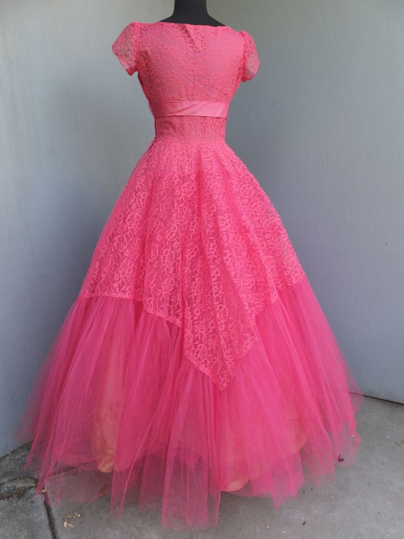May include: A pink tulle and lace gown with a fitted bodice and a full skirt. The gown has a sweetheart neckline and short sleeves. The bodice is decorated with a pink satin ribbon.