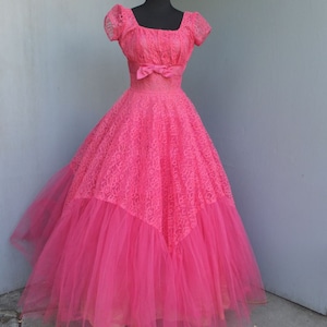 May include: A vintage pink tulle and lace ball gown with a fitted bodice and a full skirt. The dress has a square neckline, short sleeves, and a bow at the waist.
