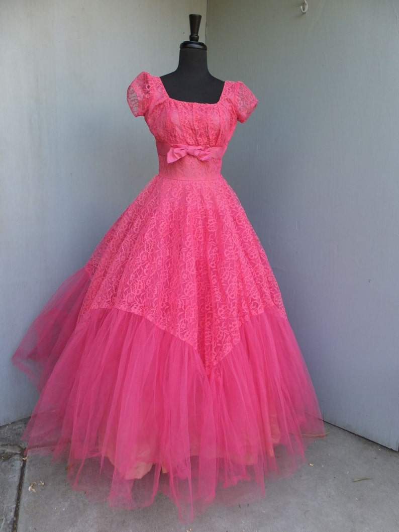 May include: A vintage pink tulle and lace ball gown with a fitted bodice and a full skirt. The dress has a square neckline, short sleeves, and a bow at the waist.