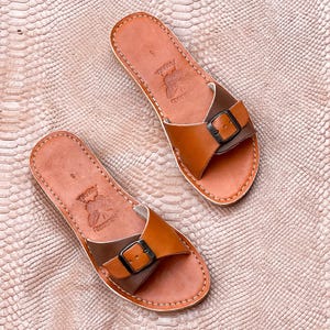 May include: A pair of brown leather sandals with a buckle closure. The sandals have a light brown footbed and a darker brown strap. The sandals are stitched with white thread and have the word "Jumana" printed on the footbed.