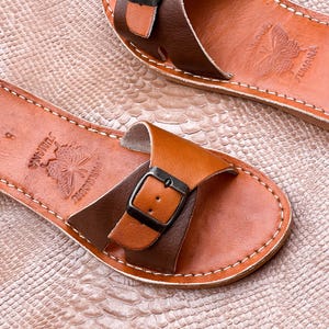 May include: A pair of leather sandals in shades of brown. The sandals feature a strap design with a buckle closure and decorative stitching. The footbed is embossed with a butterfly and the word "JUMANA".