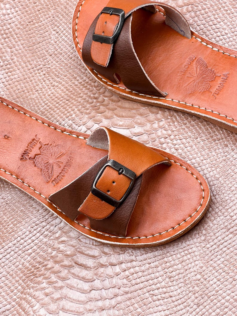 May include: A pair of leather sandals in shades of brown. The sandals feature a strap design with a buckle closure and decorative stitching. The footbed is embossed with a butterfly and the word "JUMANA".