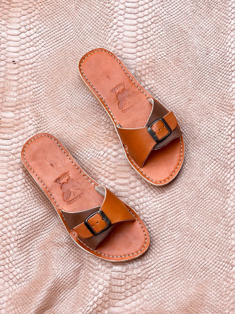 May include: A pair of brown leather sandals with a buckle closure. The sandals have a light brown footbed and a darker brown strap. The sandals are stitched with white thread and have the word "Jumana" printed on the footbed.