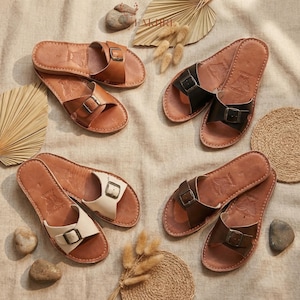 Handmade Moroccan leather buckle sandals for women in multiple colors, boho flat slide summer shoes crafted from genuine leather, minimalist artisan comfort sandals displayed in warm neutral setting