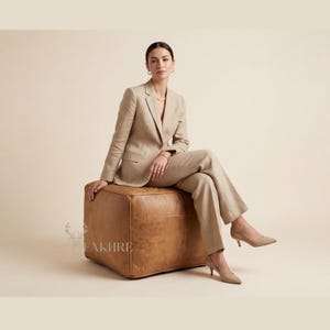Puede incluir: Una mujer con un traje color beige, sentada en un puf cuadrado de cuero marrón. El traje incluye una americana y pantalones a juego. El puf tiene un diseño cosido. La mujer lleva tacones color beige y joyas doradas.