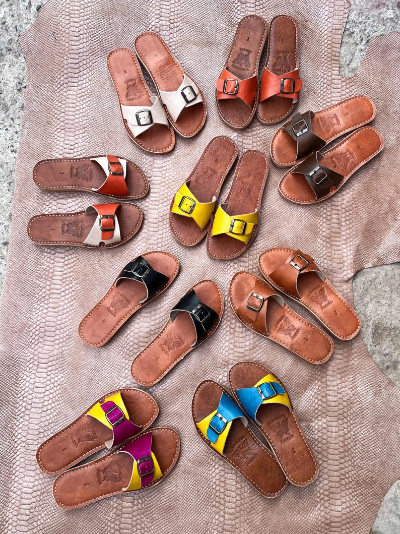 May include: A collection of leather sandals in various colors, including white, orange, yellow, black, brown, and blue. Each sandal features a buckle closure and a brown sole. The sandals are arranged on a textured, light brown surface.