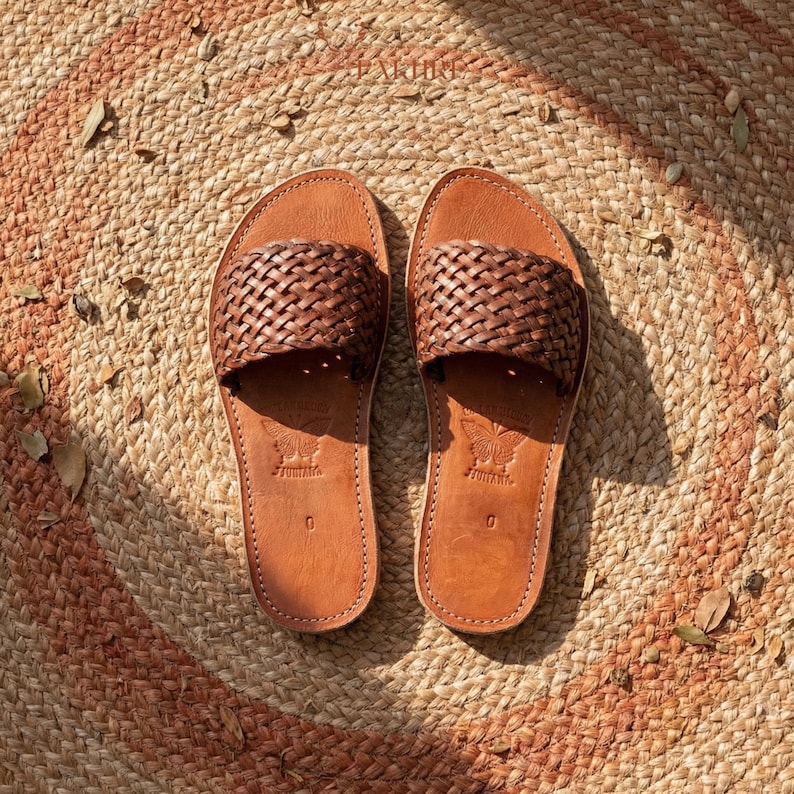 Handmade woven leather slide sandals for women placed on a natural round woven straw rug, braided genuine brown leather upper, minimalist Moroccan flat summer sandals, visible hand stitching details, warm natural sunlight, artisan craftsmanship aesthetic