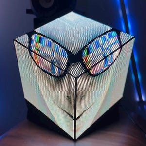May include: A cube-shaped LED display screen featuring a digital graphic of a face wearing glasses with a colourful, static-like pattern on the lenses. The screen is composed of a grid of small light-emitting diodes, creating a pixelated visual effect.