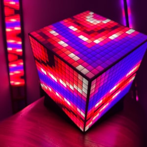 May include: A cube-shaped LED lamp with a pixelated design. The lamp features a vibrant display of red, white, and blue lights. The lamp is on a wooden surface, with a matching LED light strip in the background.