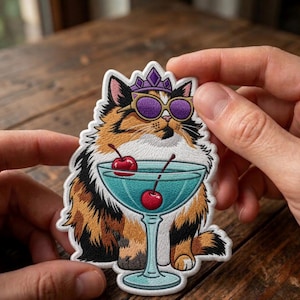 Op de afbeelding: Geborduurde patch met een lapjeskat met een paarse zonnebril en een kroon. De kat zit voor een blauwe cocktail met kersen. De patch heeft een witte rand en wordt tussen twee handen gehouden.