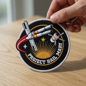 May include: Round embroidered patch with a black background. The design features a spaceship, a rising sun, and the text "PROJECT HAIL MARY." The patch has white, orange, and yellow details, and a star. The patch has a black border.