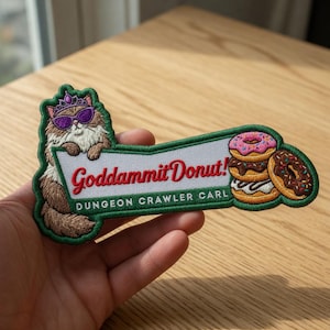Peut inclure: Écusson brodé avec un chat portant des lunettes de soleil et une couronne, tenant une bannière avec l'inscription "Goddammit Donut!" et "Dungeon Crawler Carl" en dessous. L'écusson comprend des images de beignets.