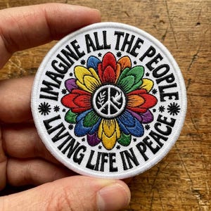 Puede incluir: Parche redondo bordado en blanco con las palabras "IMAGINE ALL THE PEOPLE LIVING LIFE IN PEACE" en negro. El parche presenta un diseño floral colorido con un símbolo de la paz en el centro. Los pétalos de la flor son de colores arcoíris.