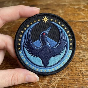 以下が含まれることがあります： 黒い背景に翼を広げた濃い青色の鳥が描かれた円形の刺繍ワッペン。デザインには、水色の三日月形、鳥の赤い目、そして小さな金色の星の縁取りが含まれています。