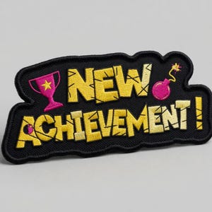 Może przedstawiać: Haftowana naszywka z napisem "NEW ACHIEVEMENT!" w kolorze żółtym, obramowanym na czarno. Naszywka przedstawia różowe trofeum, różową bombę i czarną obwódkę. Naszywka jest na szarym tle.
