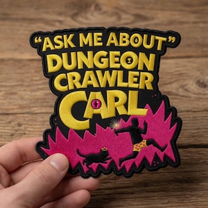 May include: Embroidered patch with the text "ASK ME ABOUT DUNGEON CRAWLER CARL" in yellow, outlined in black. The patch features a pink explosion design with a black cat wearing a crown and a figure in yellow shorts. The patch is held by a hand.