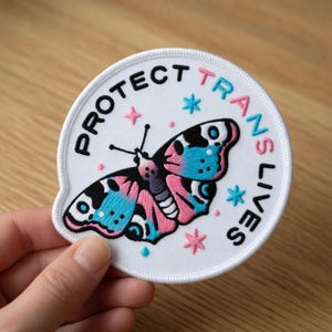 May include: A white, circular embroidered patch with the text "PROTECT TRANS LIVES" in black, pink, and blue. The patch features a butterfly with pink and blue wings, surrounded by stars. The patch is held by a hand.