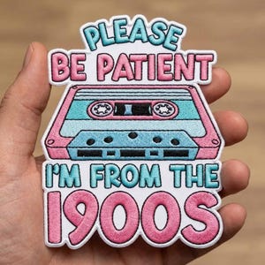 May include: Embroidered patch with the text "PLEASE BE PATIENT I'M FROM THE 1900S" featuring a retro cassette tape design in pink, blue, and white. The patch is held in a hand.
