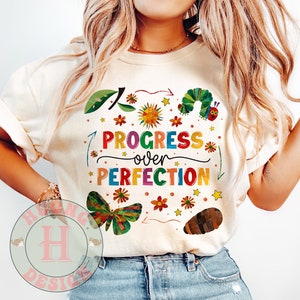 May include: A white t-shirt with a colorful graphic design featuring a caterpillar, flowers, and stars. The text "PROGRESS over PERFECTION" is written in rainbow colors.