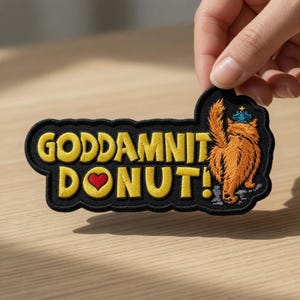 May include: Embroidered patch with the words "GODDAMNIT DONUT!" in yellow, a red heart, and a cartoon cat wearing a crown. The patch has a black border and is held by a hand.
