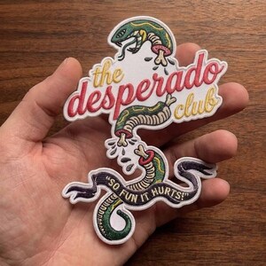 Peut inclure: Écusson brodé avec un motif de serpent vert et jaune et le texte "the desperado club" en rouge et jaune. L'écusson comporte également la phrase "SO FUN IT HURTS!" sur une bannière noire.