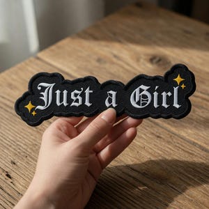 Peut inclure: Écusson brodé noir avec l'inscription "Just a Girl" en lettres gothiques blanches. Le patch a une bordure noire en forme de nuage et des étoiles jaunes. Le patch est posé sur une surface en bois.